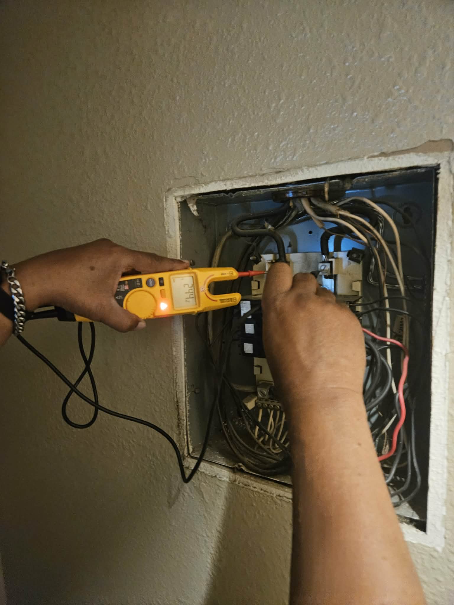 Hands using a yellow digital multimeter to test voltage in an open electrical breaker box.