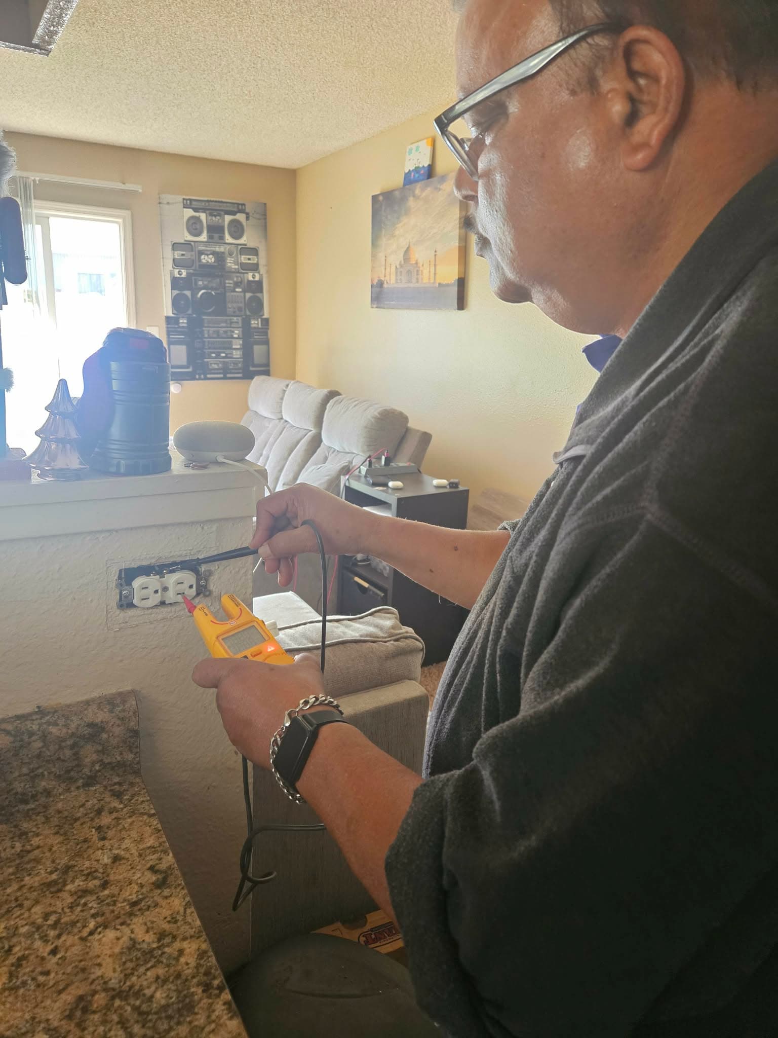Man using a yellow digital multimeter to test an exposed electrical wall outlet indoors.