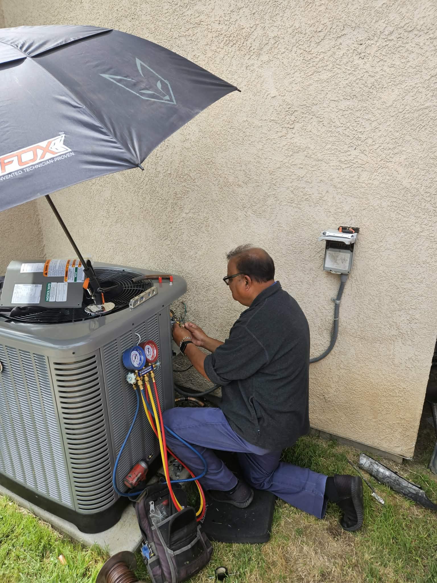 HVAC technician kneeling while repairing an outdoor air conditioning unit under a black work umbrella.