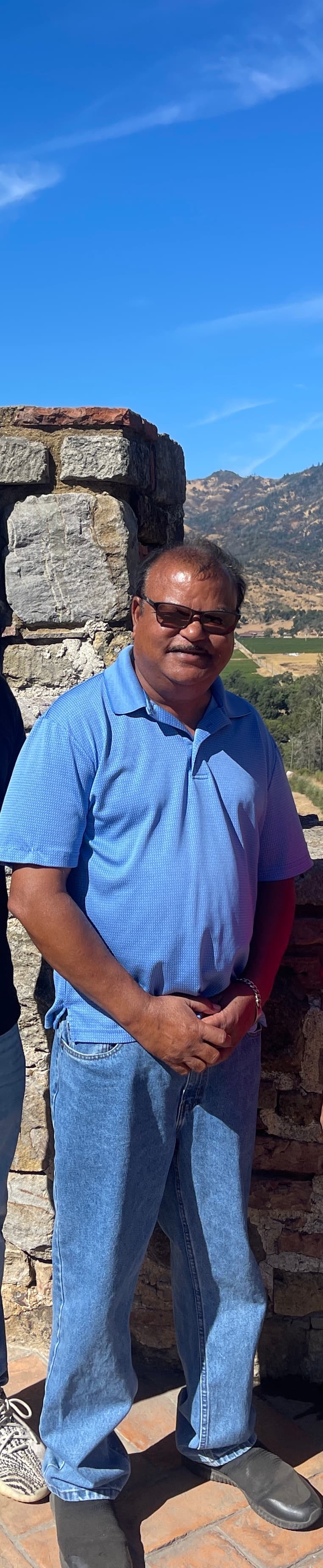 Man in blue polo and sunglasses stands by a stone wall with scenic mountain views.