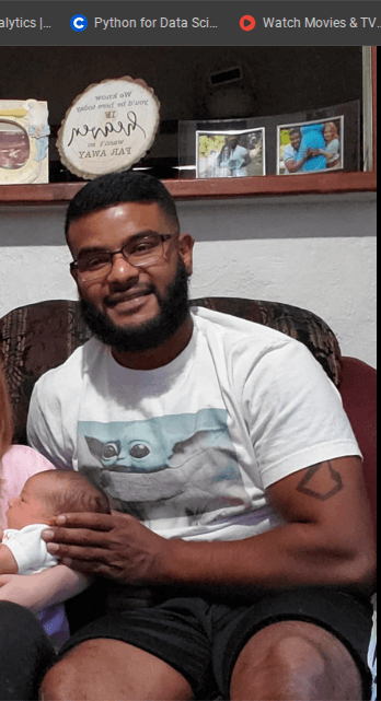 Bearded man in a Grogu t-shirt sits and gently holds a newborn baby indoors.