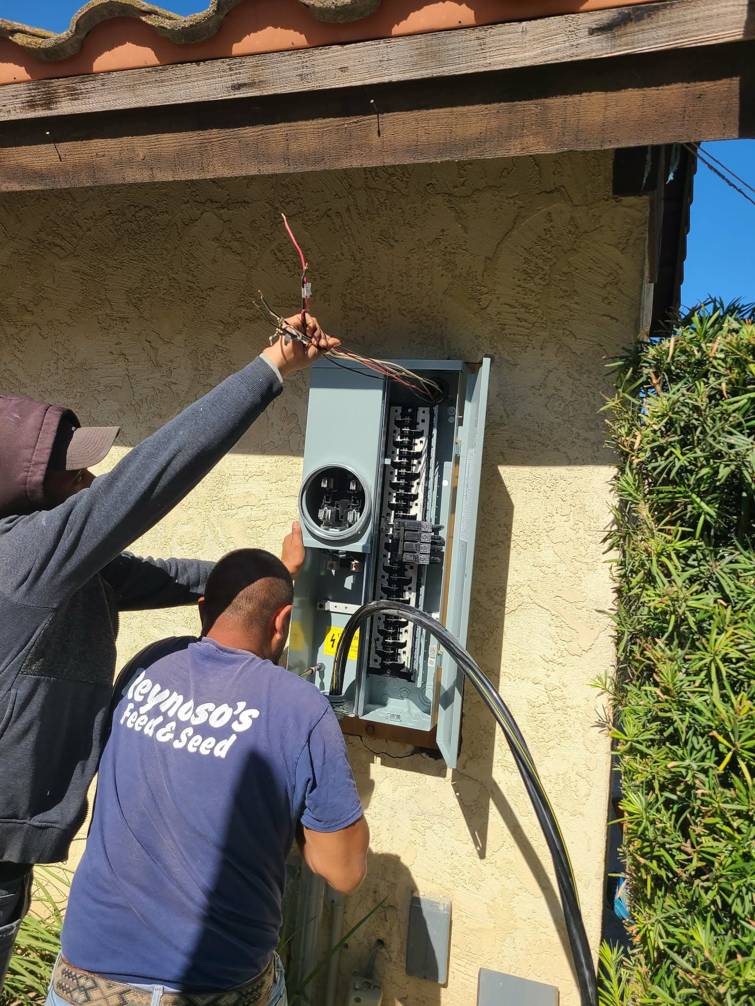Two workers installing wiring into an open outdoor electrical service panel on a stucco wall.