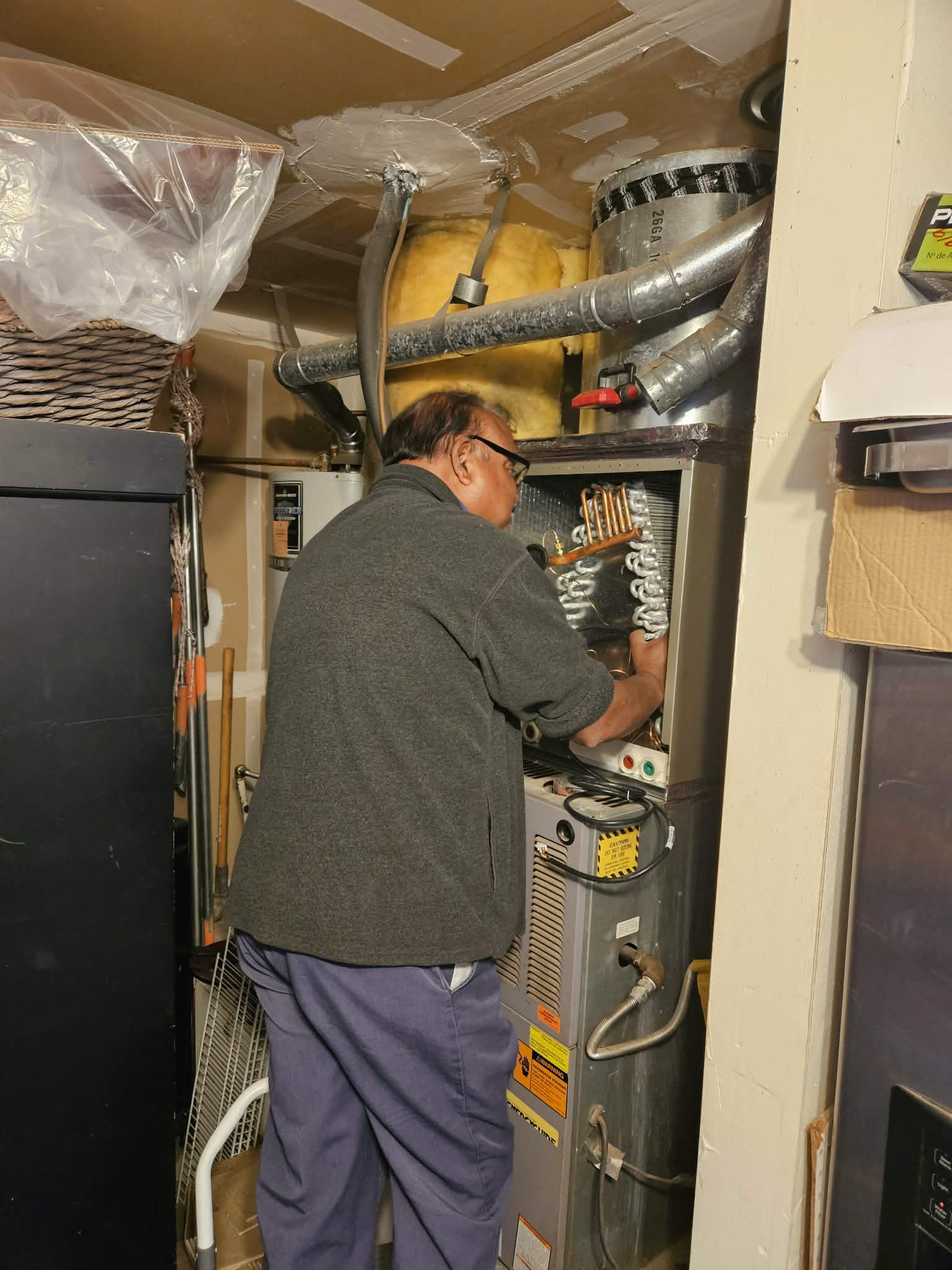 A man in a grey fleece repairs an open HVAC unit in a cluttered utility room.
