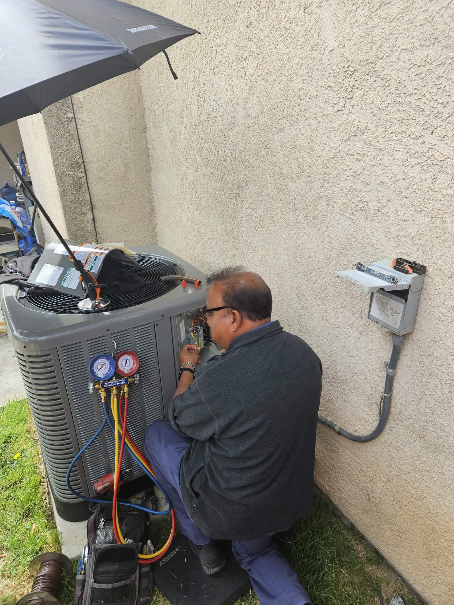 HVAC technician repairs an outdoor air conditioning unit with pressure gauges under a black umbrella.
