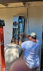 Two men working on an open outdoor electrical panel with various wires and components visible.